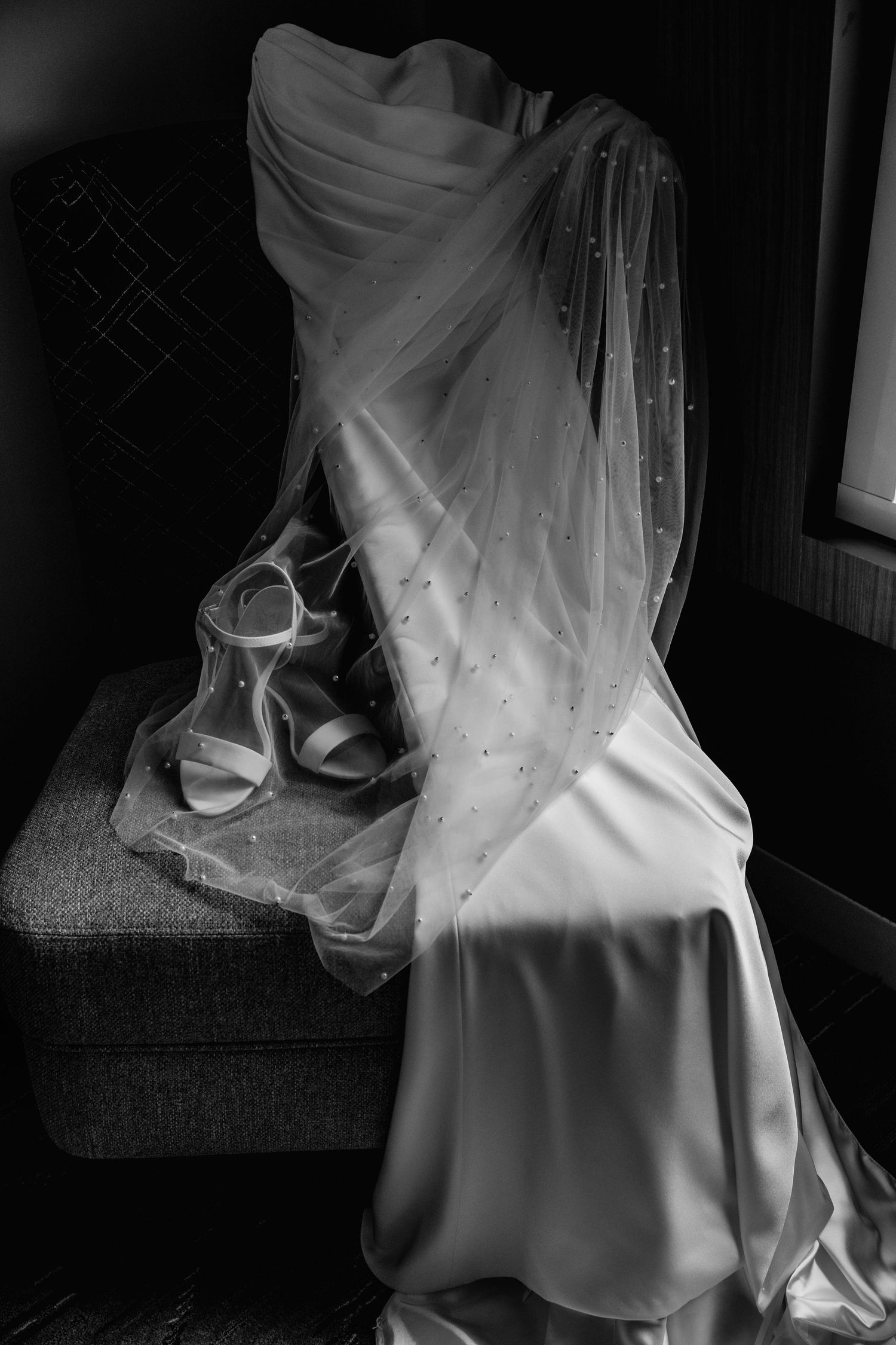A delicate white wedding veil with tulle fabric cascades elegantly over a formal dress in black and white photography.