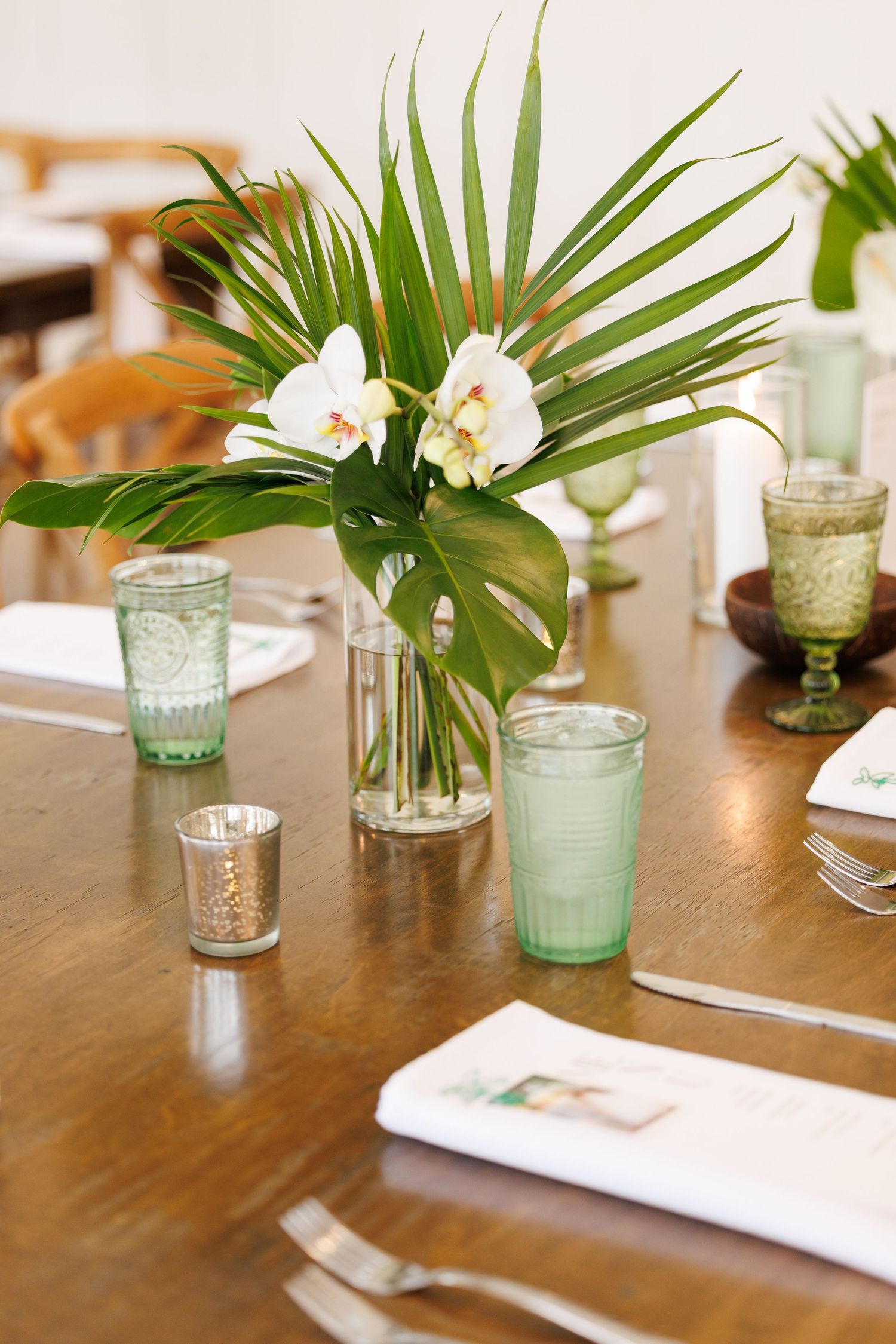 Elegant table setting with tropical palm leaves and white orchids in seafoam green glass vases create a modern coastal vibe at Bella Cavalli