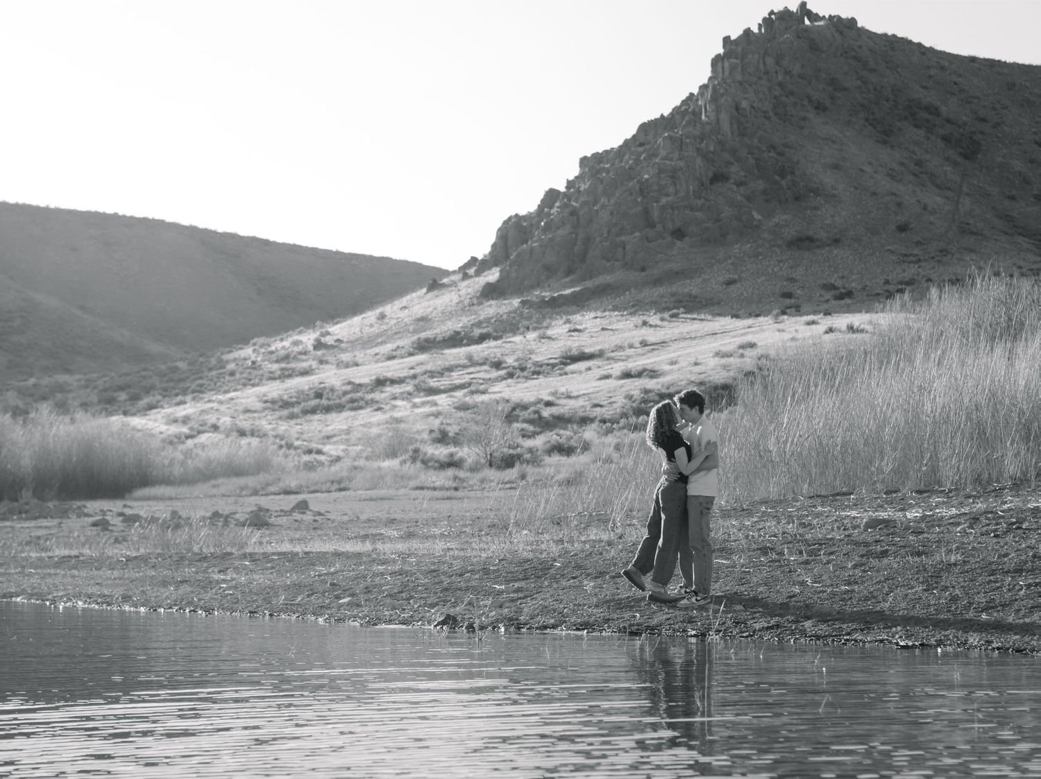 Black-and-white photo of an engaged couple standing close together near the water's edge with a large hill or mountain in the background.