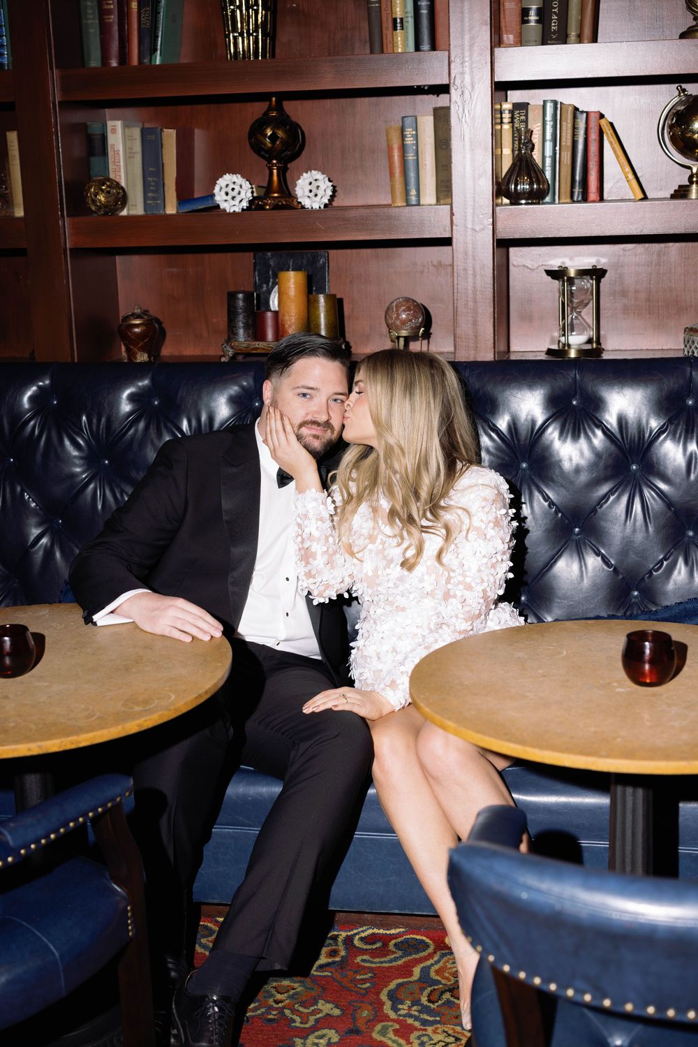 Romantic couple in formal attire sharing an intimate moment on a tufted leather banquette in an elegant library setting.