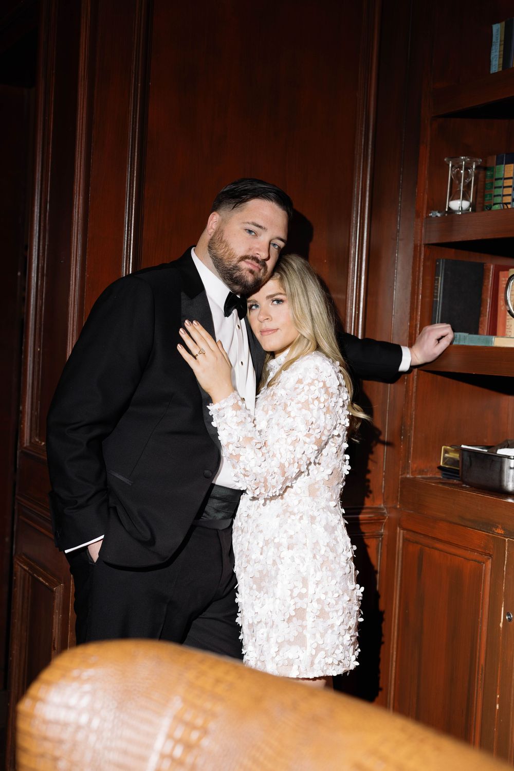 A glamorous couple poses in formal attire against wooden bookshelves in an elegant library setting.