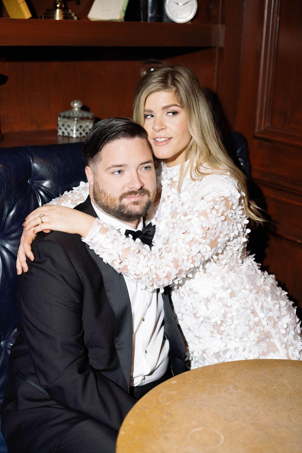 A couple in formal attire poses together on a leather couch in an elegant study setting.