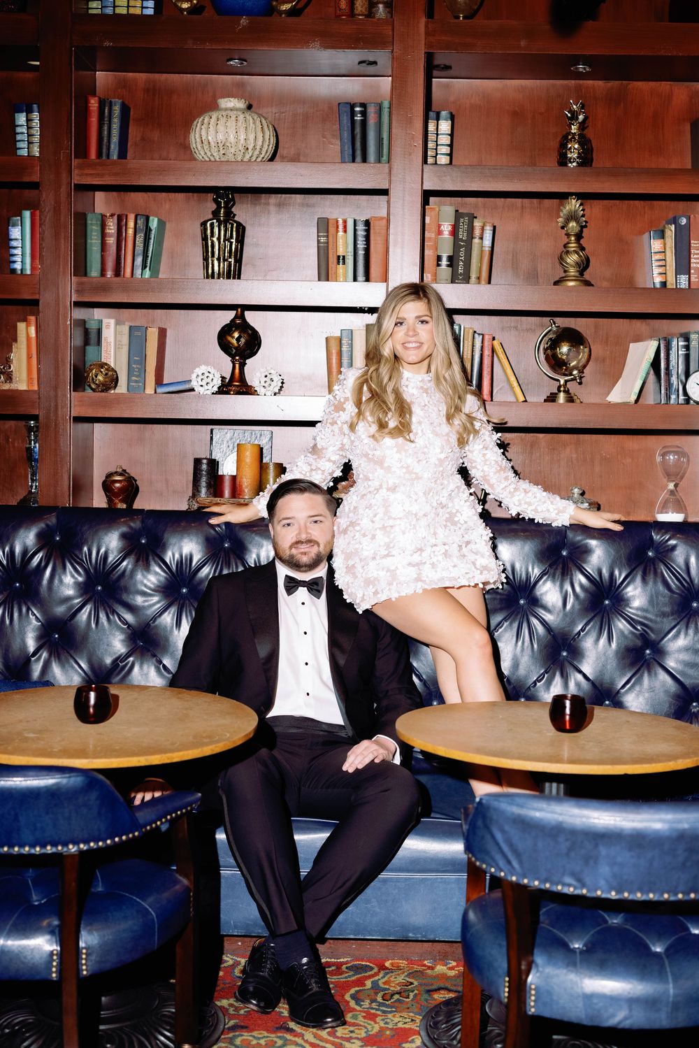 A couple poses together in a luxurious library setting with blue tufted leather chairs and wooden bookshelves.