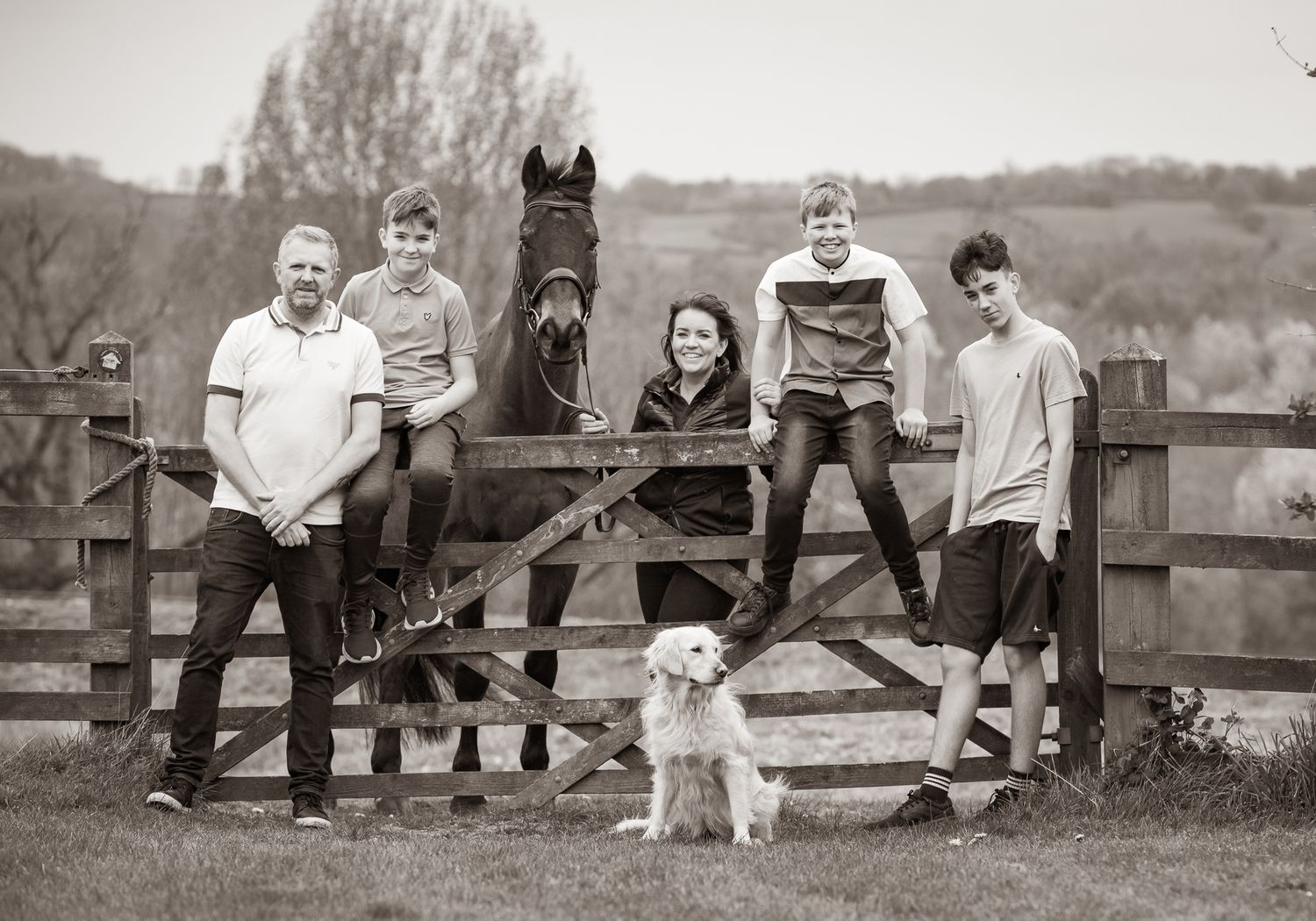 Black and white family photo on a rustic farm with a black horse and white dog by a wooden fence.
