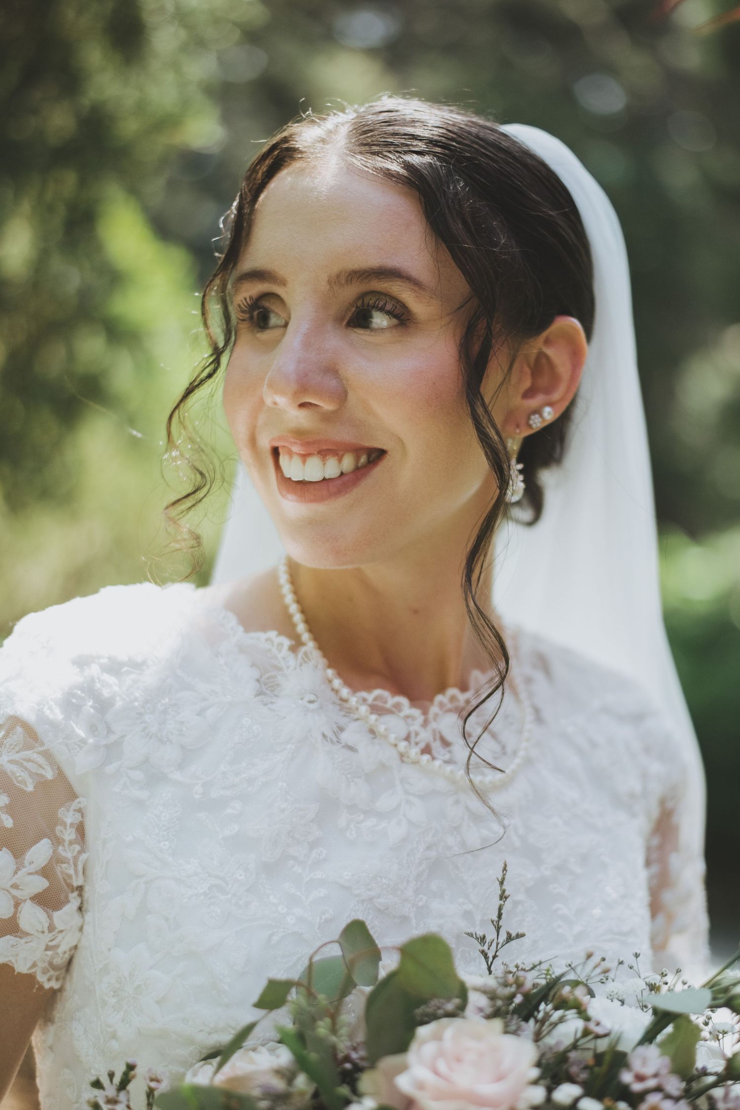 A bride in a white lace dress with long veil smiles while holding a delicate pink and green bouquet in outdoor setting.