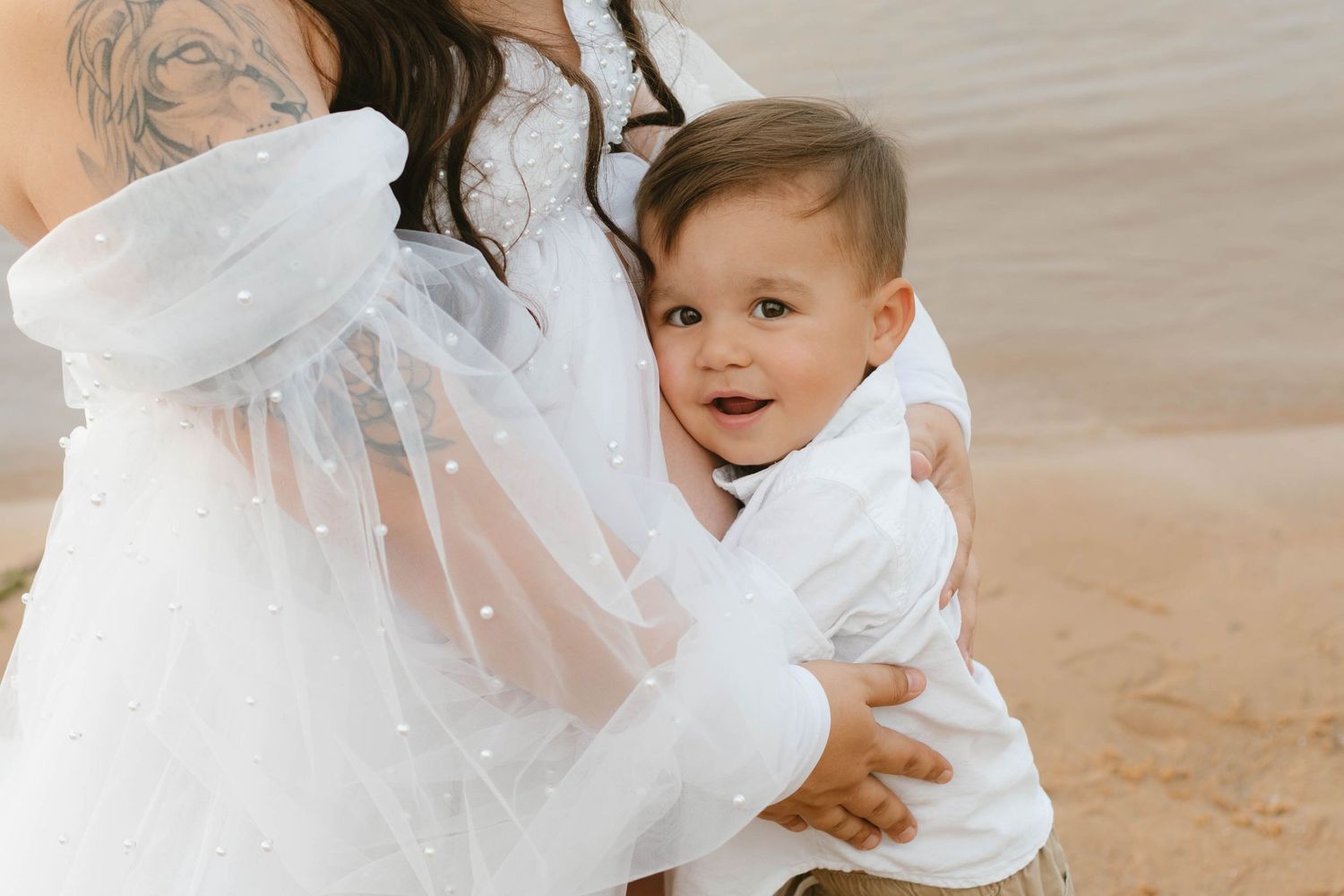 Pregnant woman in white flowing maternity dress with pearl details and long sleeves cradles her baby bump.