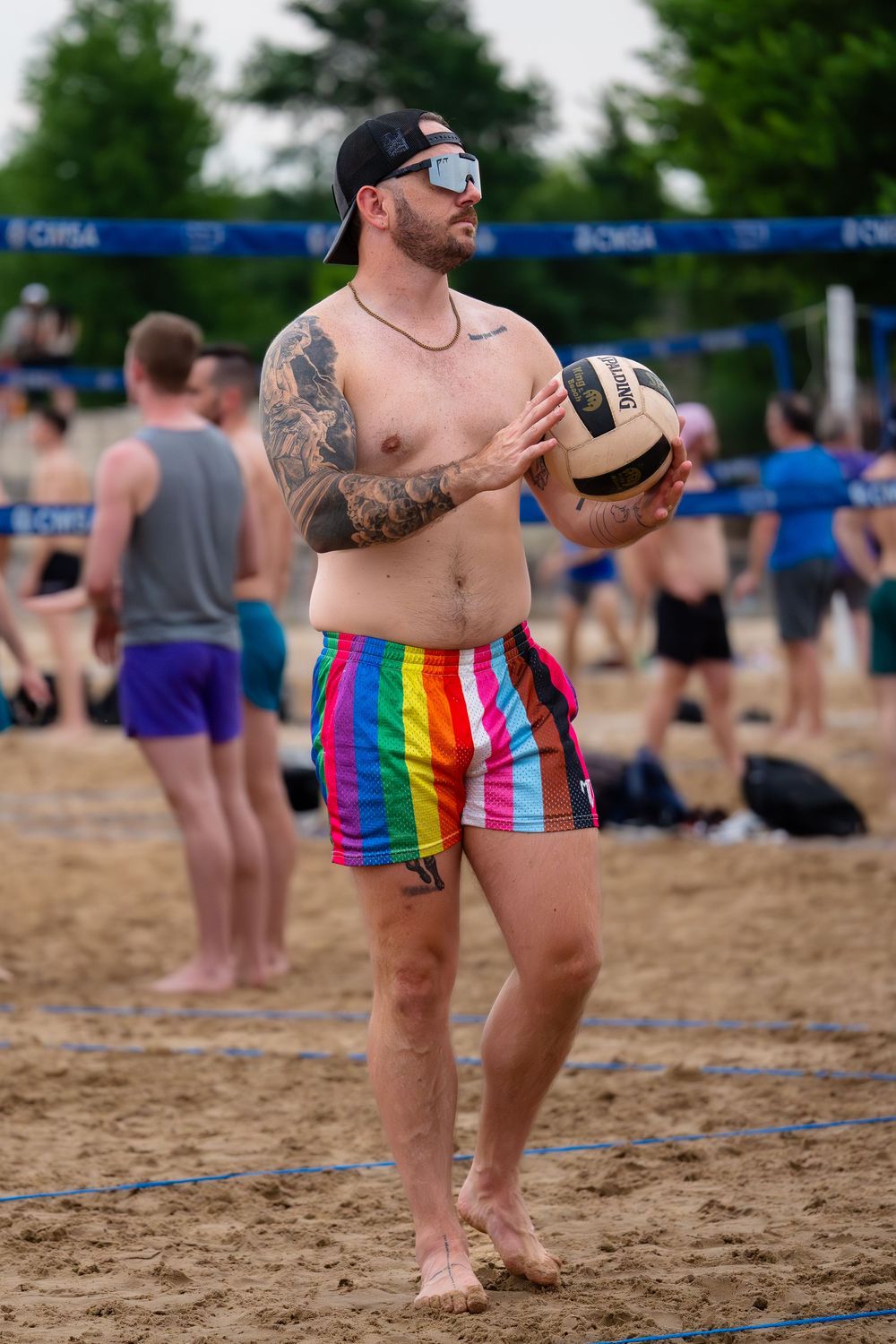A shirtless athlete waits for the opportunity to serve the volleyball over the net in multicolor shorts