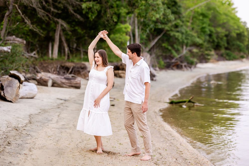A couple in white outfits dance together on a beach shoreline with trees and driftwood in the background.