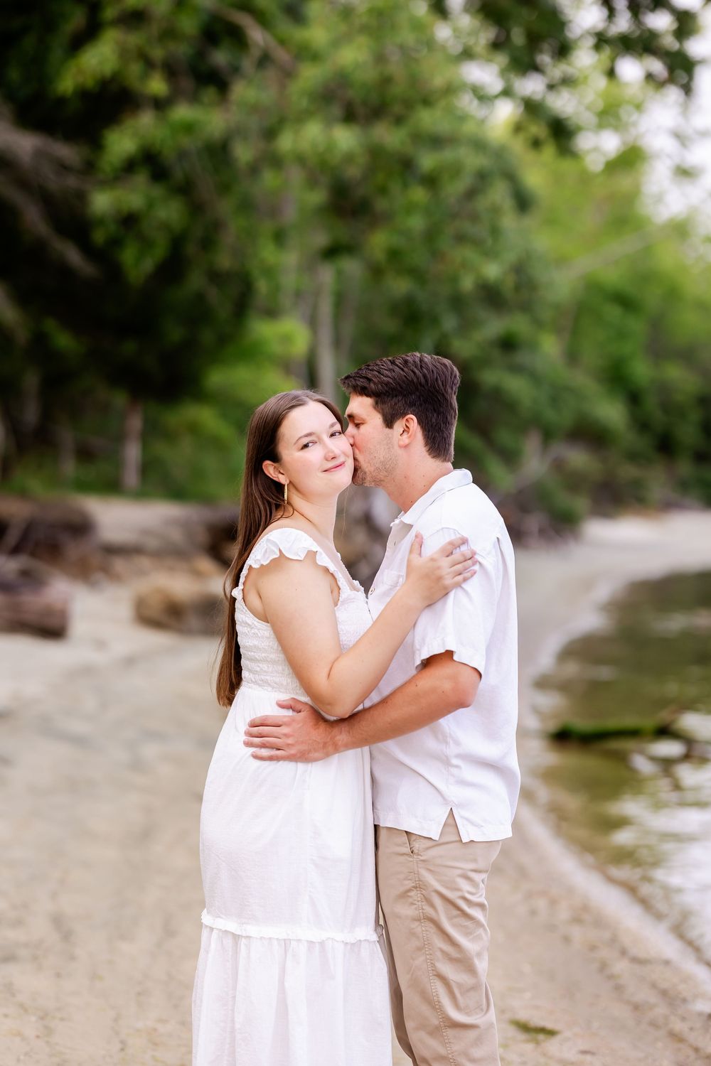 A romantic couple embraces on a sandy beach with trees and water in the background.
