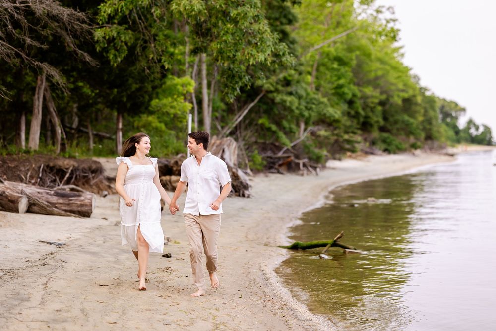 Two people stroll together barefoot along a peaceful sandy beach with trees lining the shoreline.