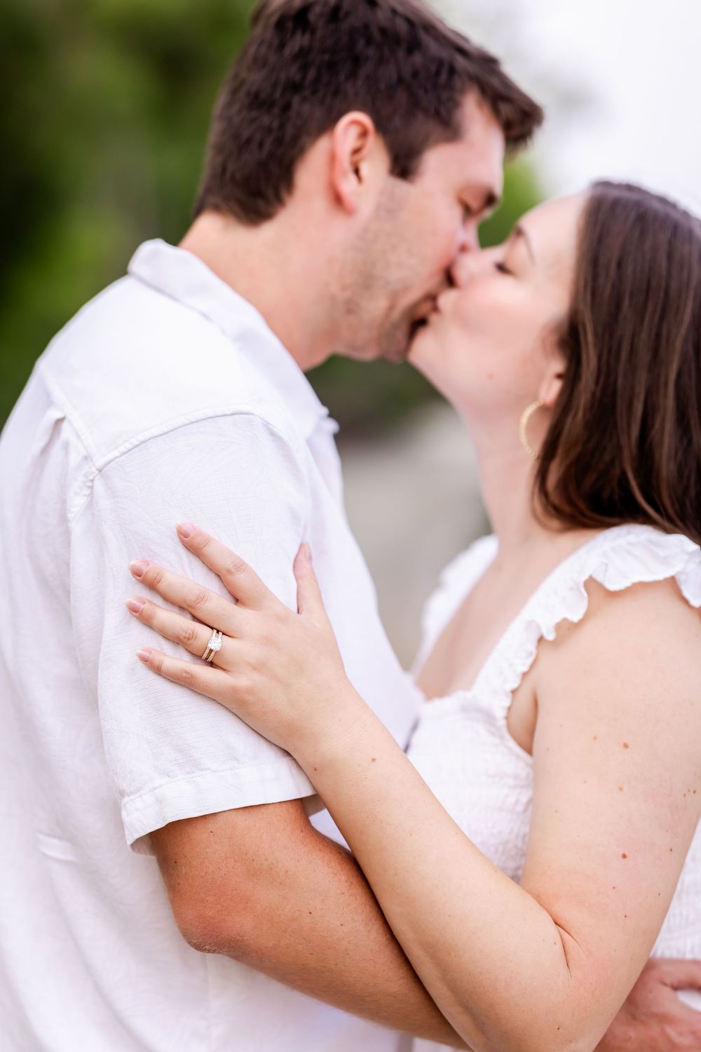 A tender romantic moment between a couple sharing a kiss outdoors, with the woman wearing white and displaying her ring.