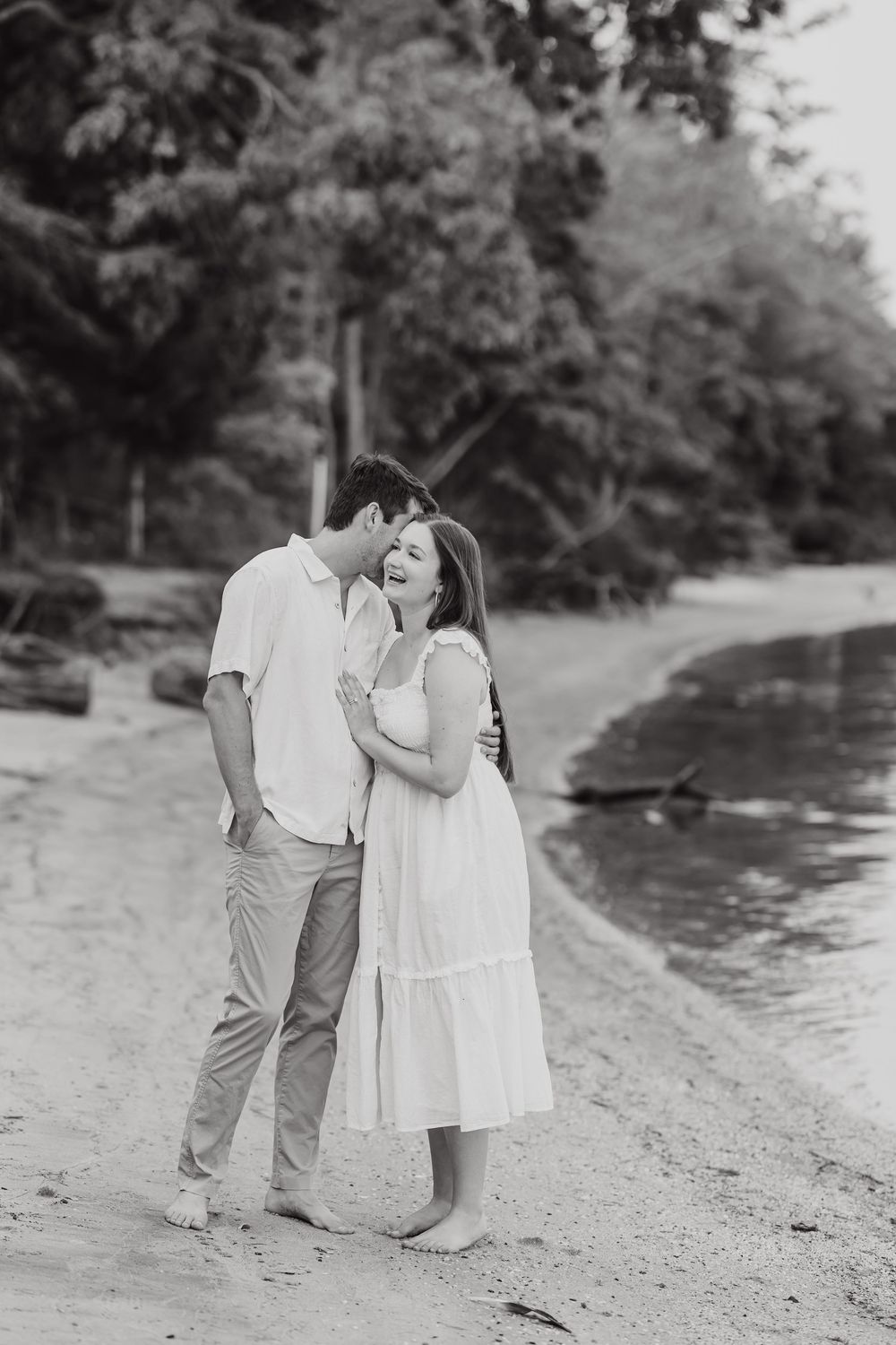 A romantic black and white photograph of a couple sharing an intimate moment on a lakeside beach.