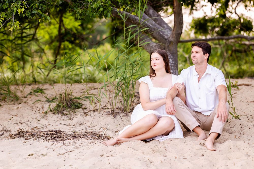 A couple sits together on a sandy beach near trees on a sunny summer day.