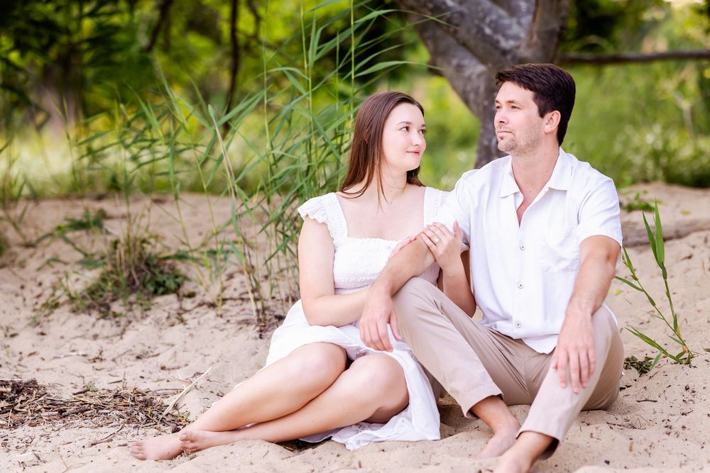A couple wearing white clothing sits together on a sandy beach during a romantic outdoor photography session.