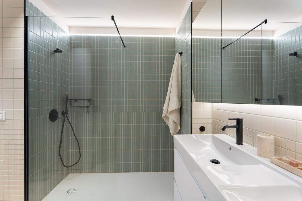 Modern bathroom interior with sage green tile, glass shower enclosure, white countertop and minimalist fixtures.