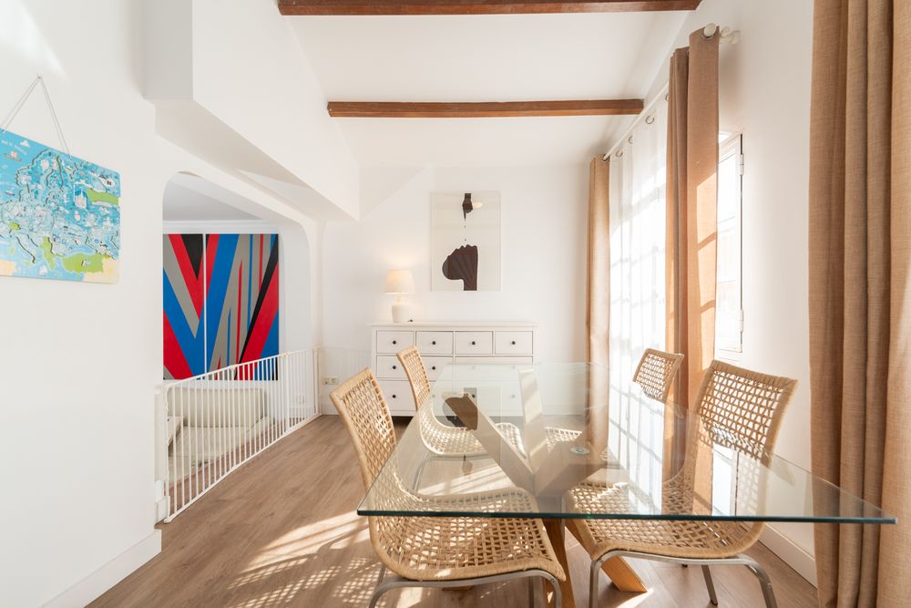 Modern dining area with glass table, woven chairs, wood beams, and colorful wall art in a bright white interior space.