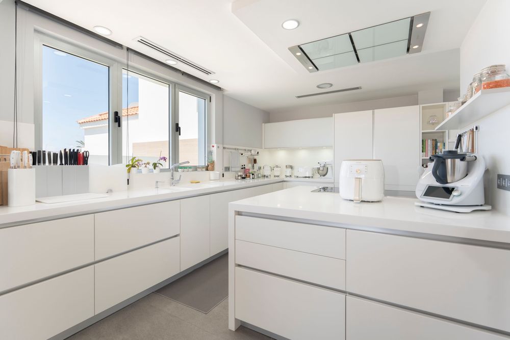Modern white kitchen interior with sleek handleless cabinets, quartz countertops and bright natural light from large window.