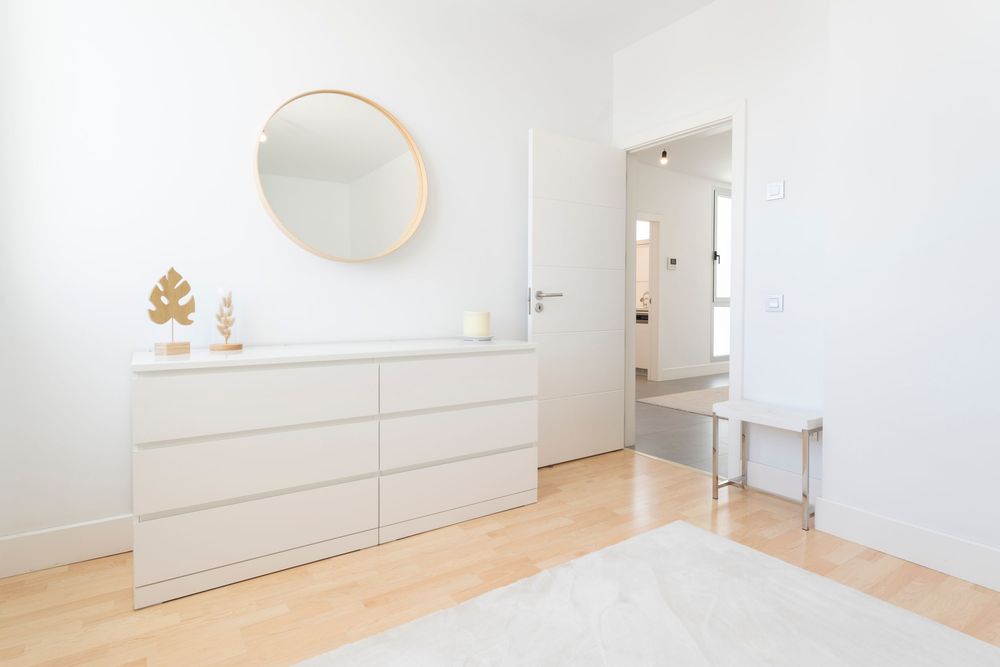Minimalist bedroom storage featuring a white dresser, round mirror, and light wooden flooring.