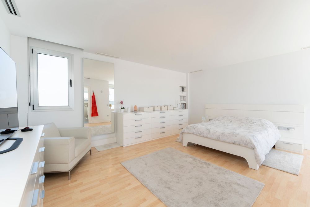 Spacious white bedroom with modern furniture, hardwood floors, and natural light from large window.