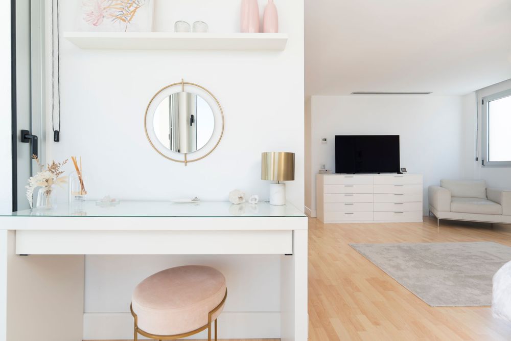 Modern vanity area with round mirror, pink stool, and white furniture in bright bedroom interior.