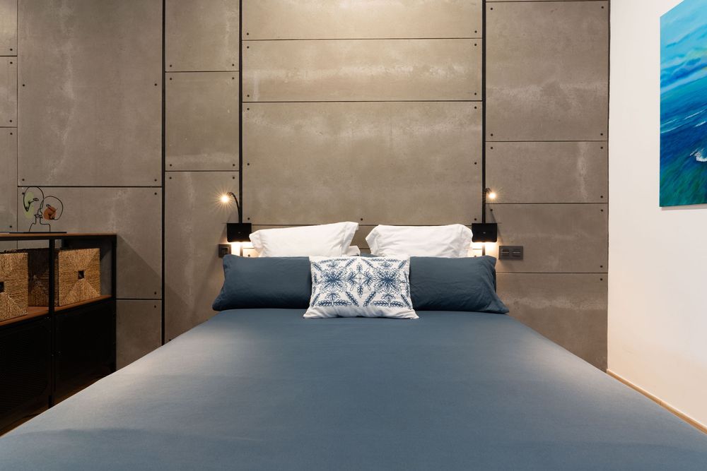 Modern bedroom interior with gray bed, concrete wall panels, and blue ocean artwork mounted beside the bed.