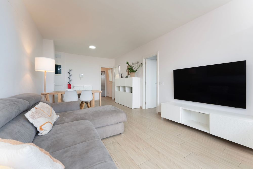 Modern minimalist living room with gray sectional sofa, wall-mounted TV, and light wood flooring in open concept space.