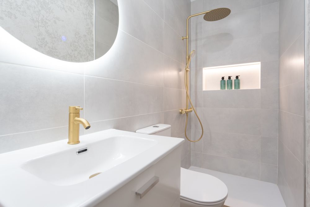Modern minimalist bathroom with white fixtures, gold faucet, and illuminated round mirror above floating sink.