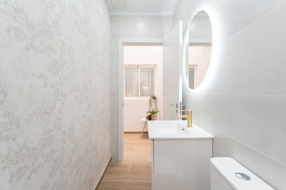 Bright white bathroom corridor with round mirror, floating vanity and floral wallpaper accent wall.