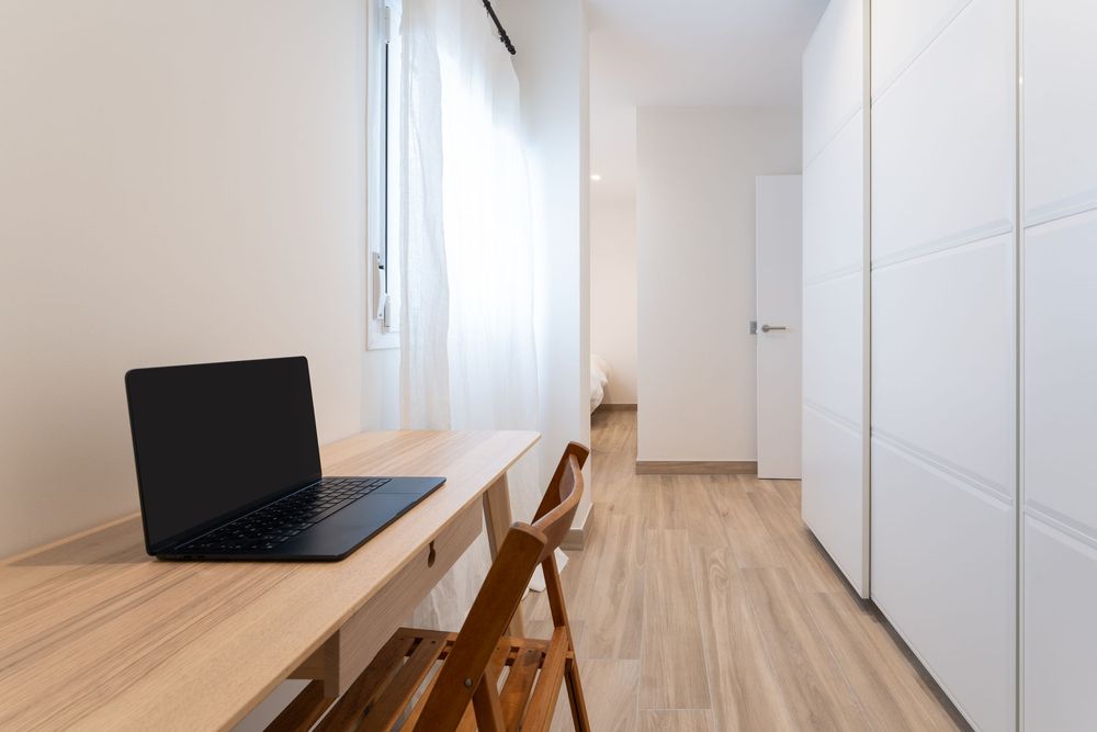 Minimalist home office setup with laptop on wooden desk, white walls, and built-in storage in narrow hallway.