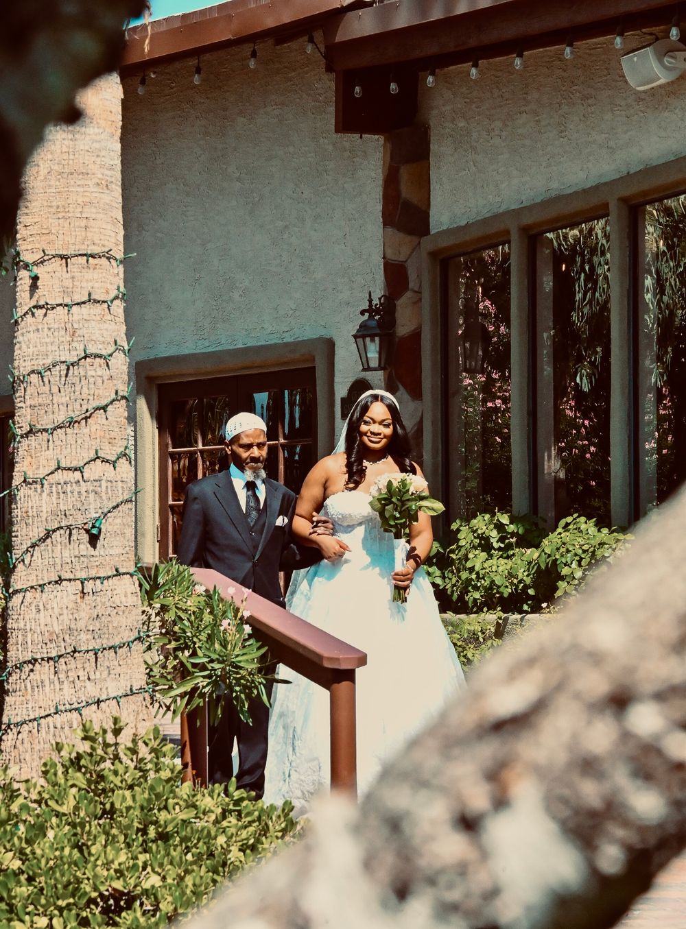 Bride in white gown holding bouquet walks with escort from venue entrance on sunny wedding day.
