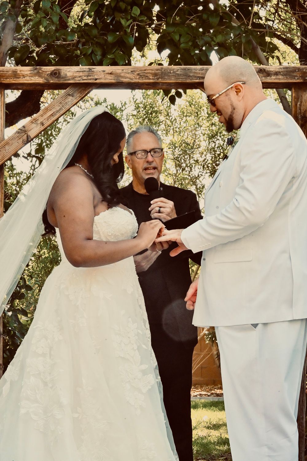 Bride placing wedding ring on groom's finger during outdoor ceremony under rustic wooden arch with officiant.