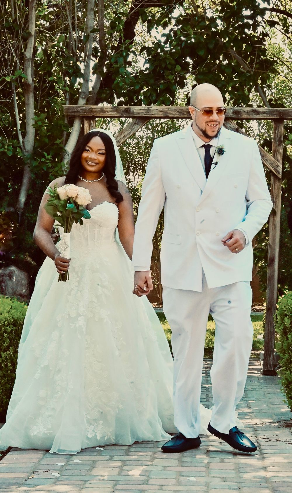 A bride in a white lace gown holding flowers walks hand-in-hand with a groom in a white suit at an outdoor wedding.