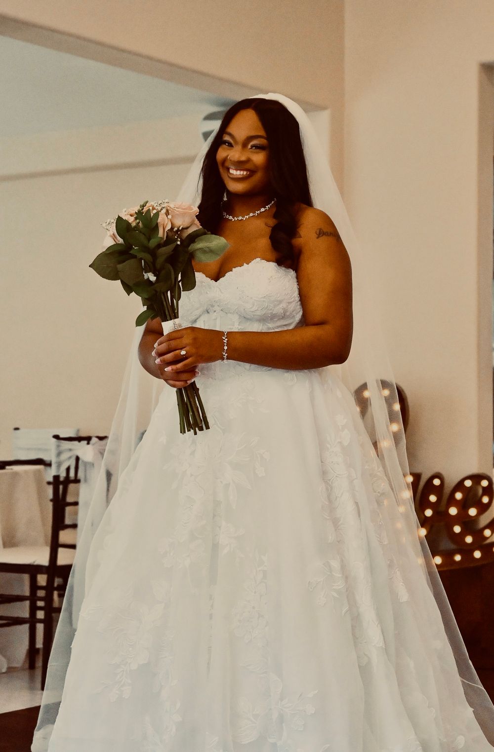 Smiling bride in a white strapless lace ball gown with veil holds a pink rose bouquet at her wedding ceremony.