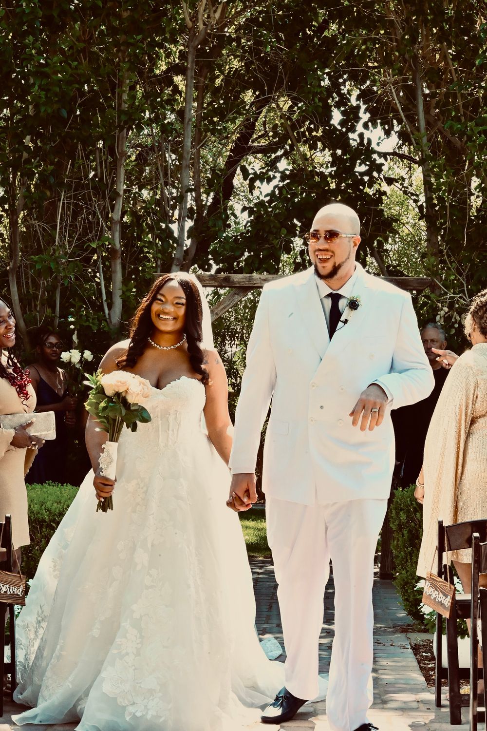 Bride in lace gown and groom in white suit walk down outdoor wedding aisle holding hands amid lush greenery.