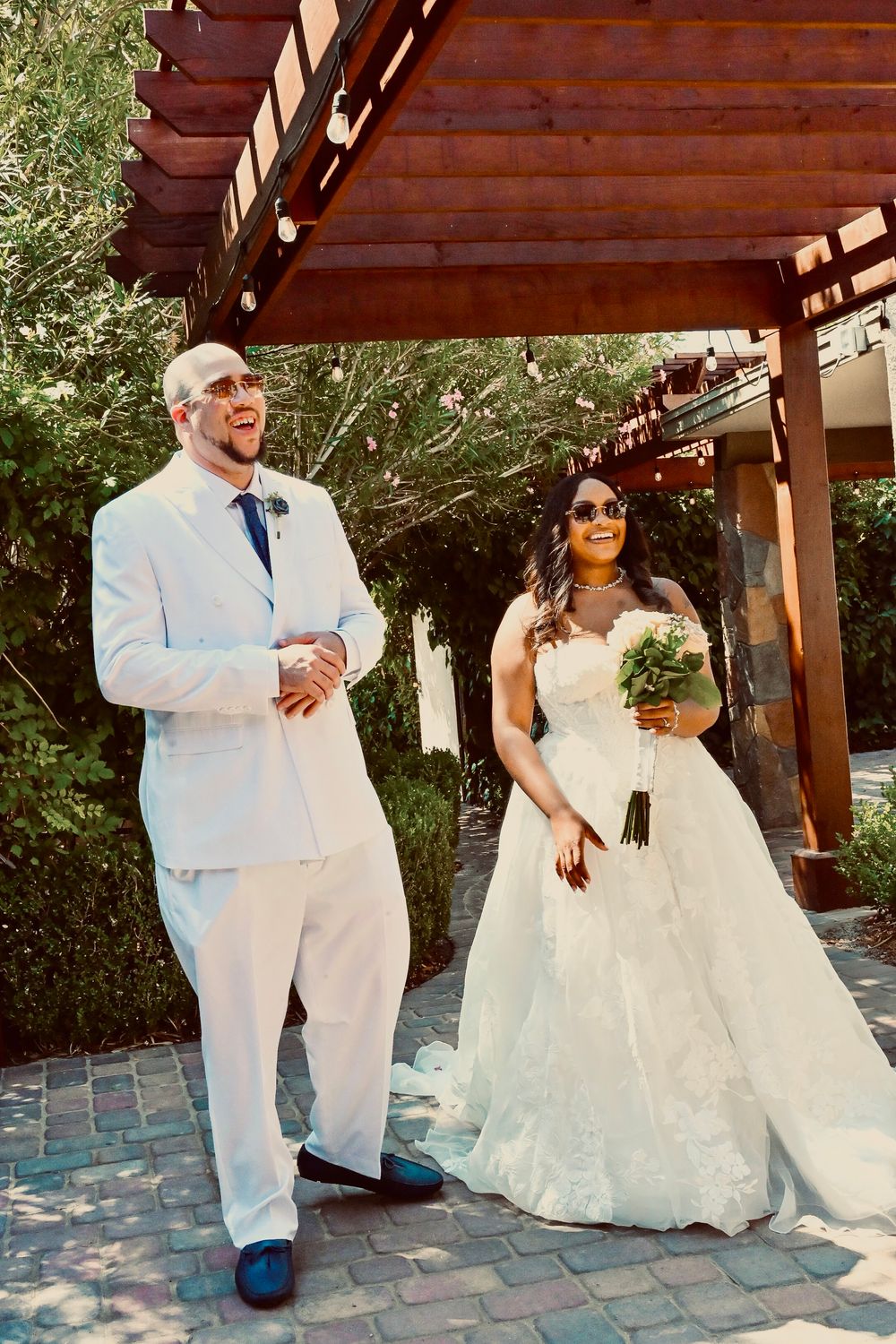 Bride and groom laughing together outdoors under wooden pergola, bride holding green bouquet in white lace ballgown.