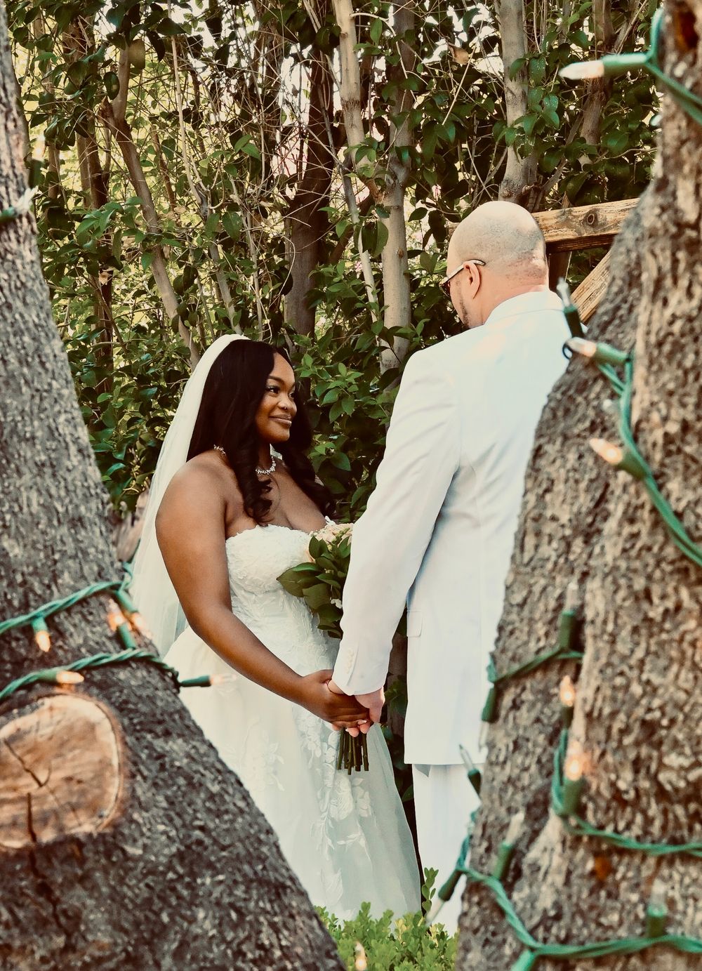 Bride and groom holding hands at outdoor woodland wedding ceremony surrounded by trees and lights.