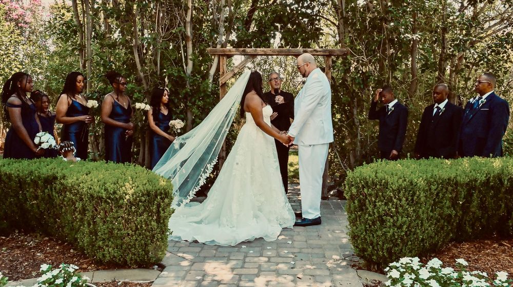 Outdoor garden wedding ceremony with bride in white gown and veil, groom in white suit, under wooden arch with guests.