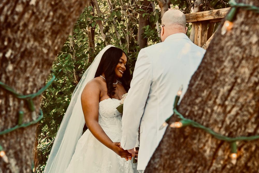 Bride in white gown and veil holds hands with groom in gray suit during outdoor wedding ceremony among trees.