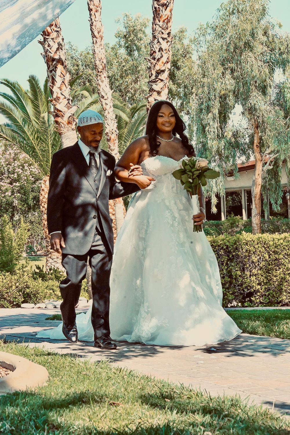 Bride in white gown walks arm-in-arm with suited escort along palm-lined outdoor path on sunny wedding day.