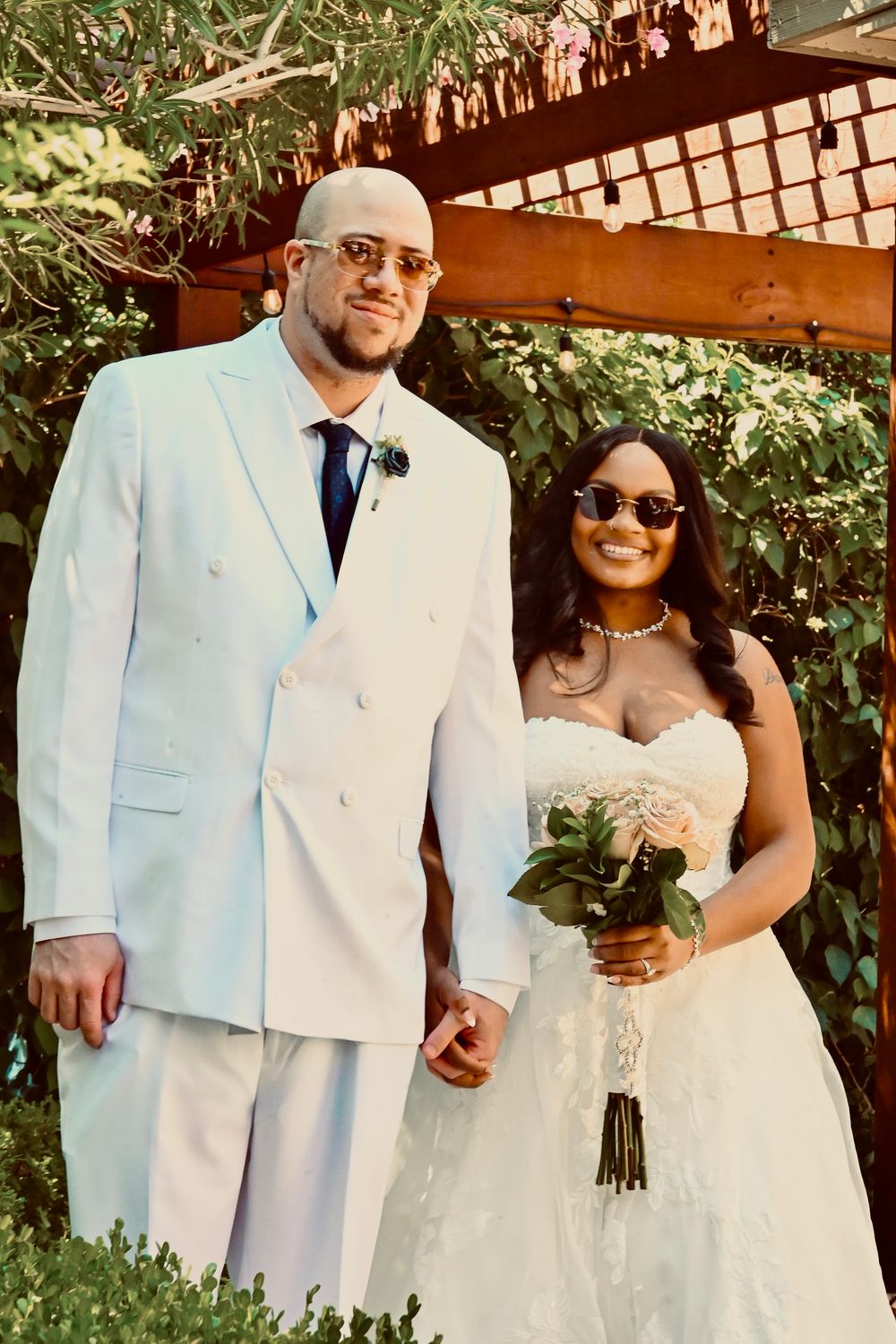 Interracial couple posing outdoors on wedding day, groom in white suit and bride in strapless gown holding bouquet.