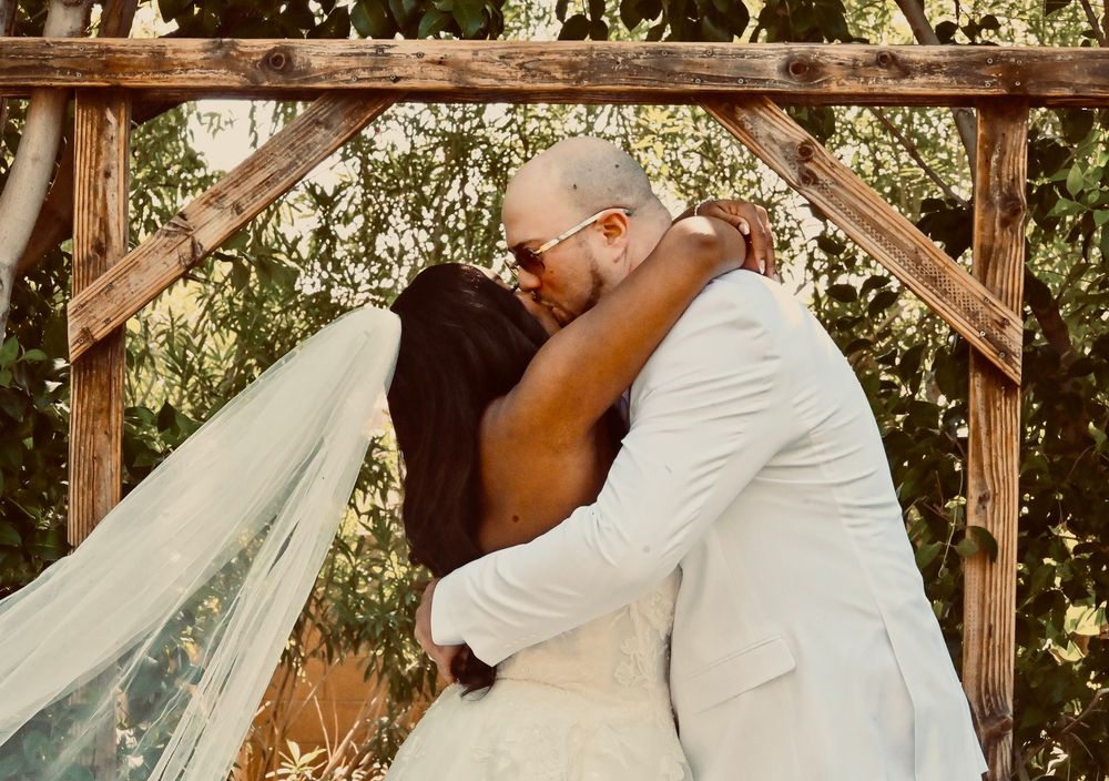 Newlyweds sharing first kiss under rustic wooden arch surrounded by lush greenery at outdoor wedding.