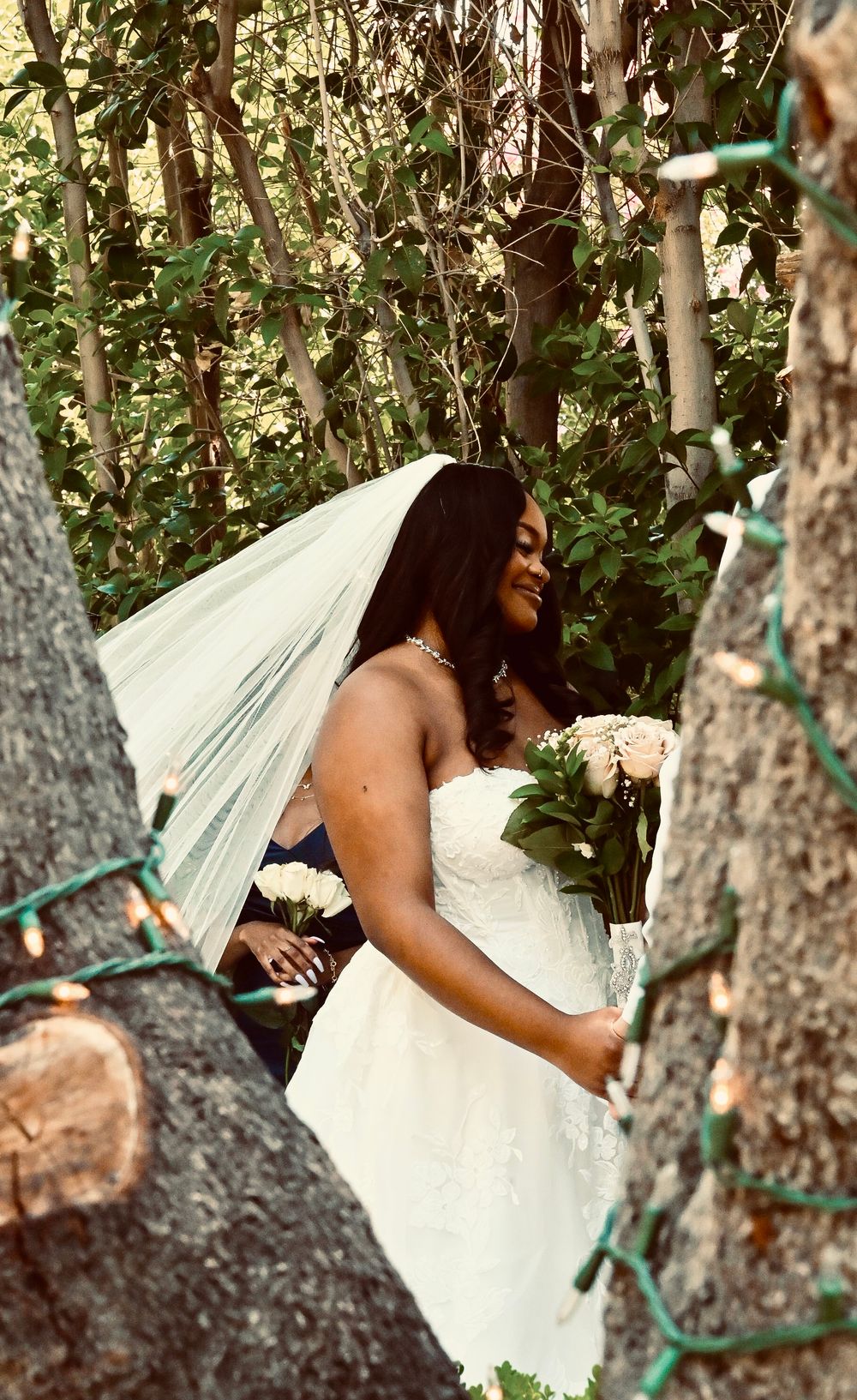 Bride in strapless white gown and veil holds bouquet of roses while standing among trees with string lights.
