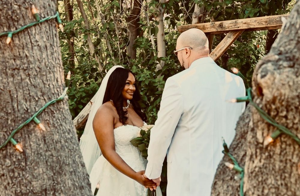 Bride in white strapless gown and veil smiles at groom during intimate outdoor woodland wedding ceremony with string lights.