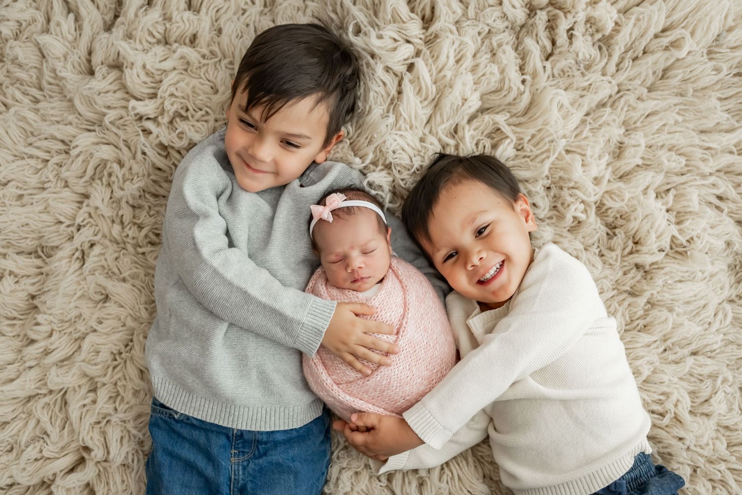 Five heartwarming photos of siblings cuddling together with a baby on a cream-colored plush rug.