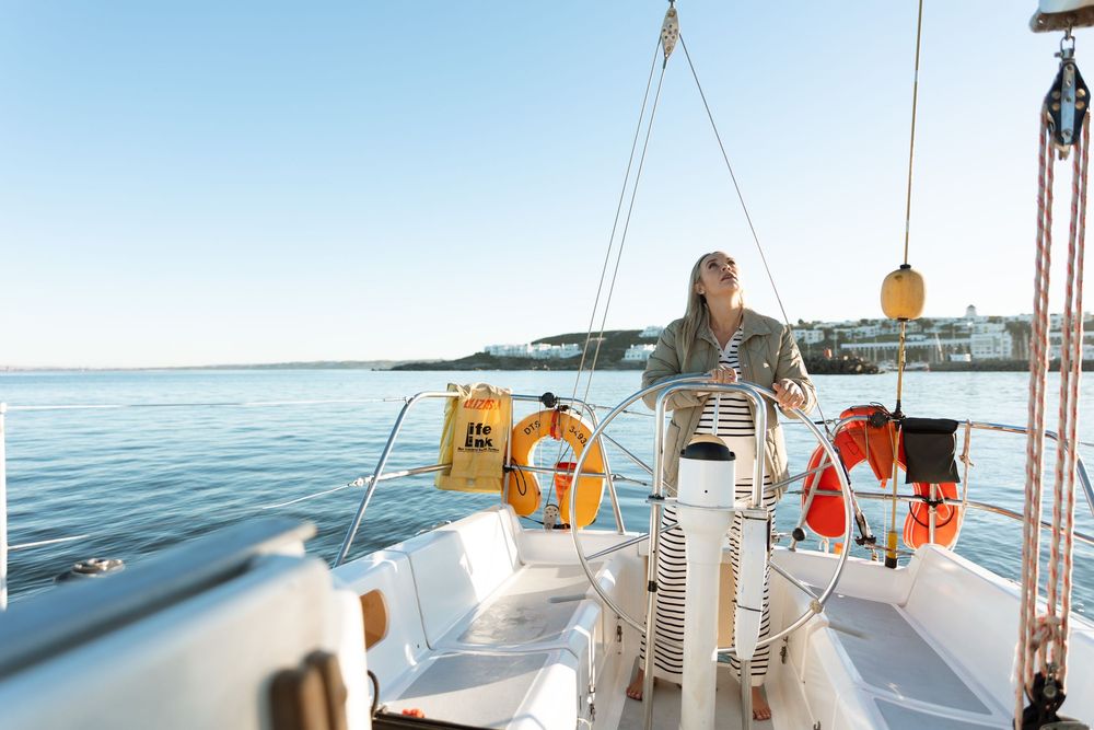 Person steering a sailboat on calm blue waters near a coastal shoreline on a sunny day.