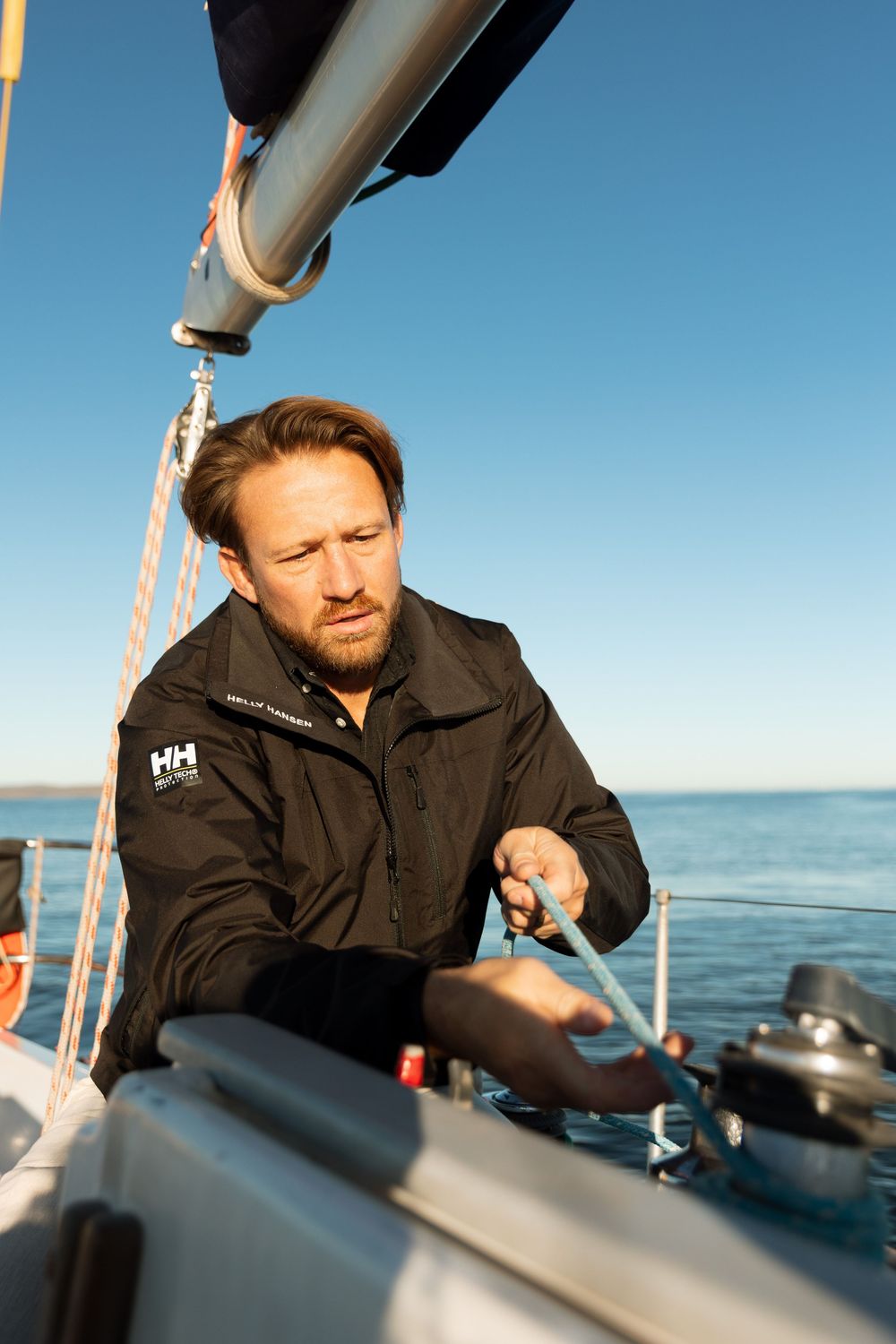 Person in black jacket adjusts equipment on sailboat deck against blue sky and ocean background.