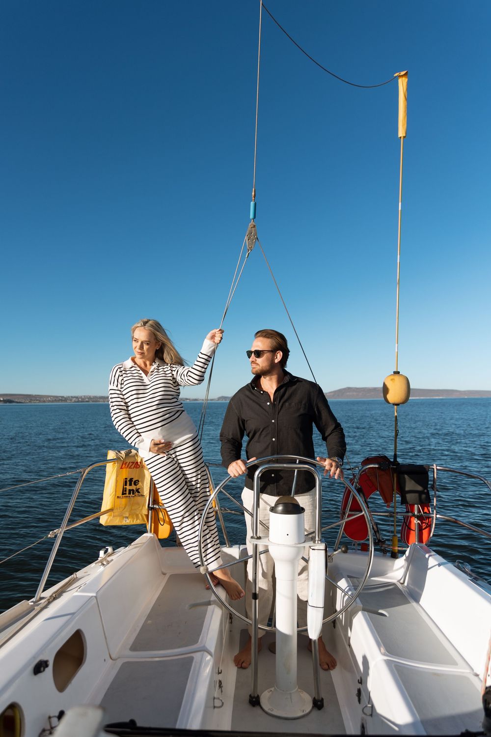 A couple navigates their sailboat on calm waters under bright blue skies.