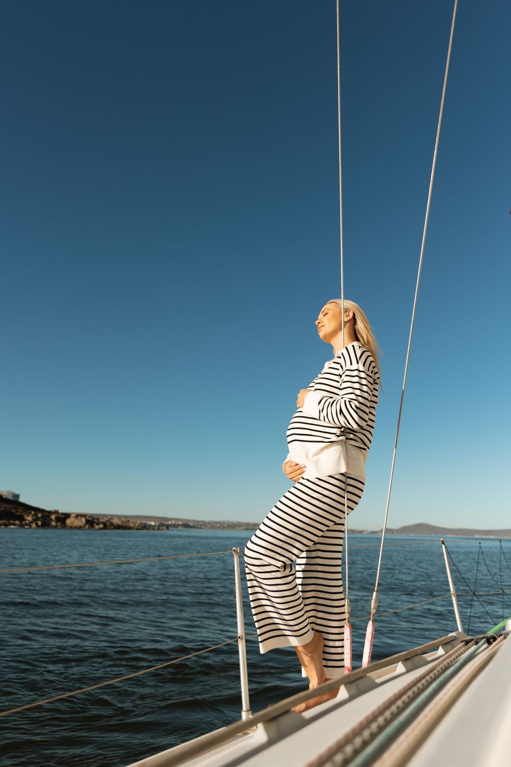 A figure in striped clothing sits on the bow of a sailboat against a blue ocean horizon.