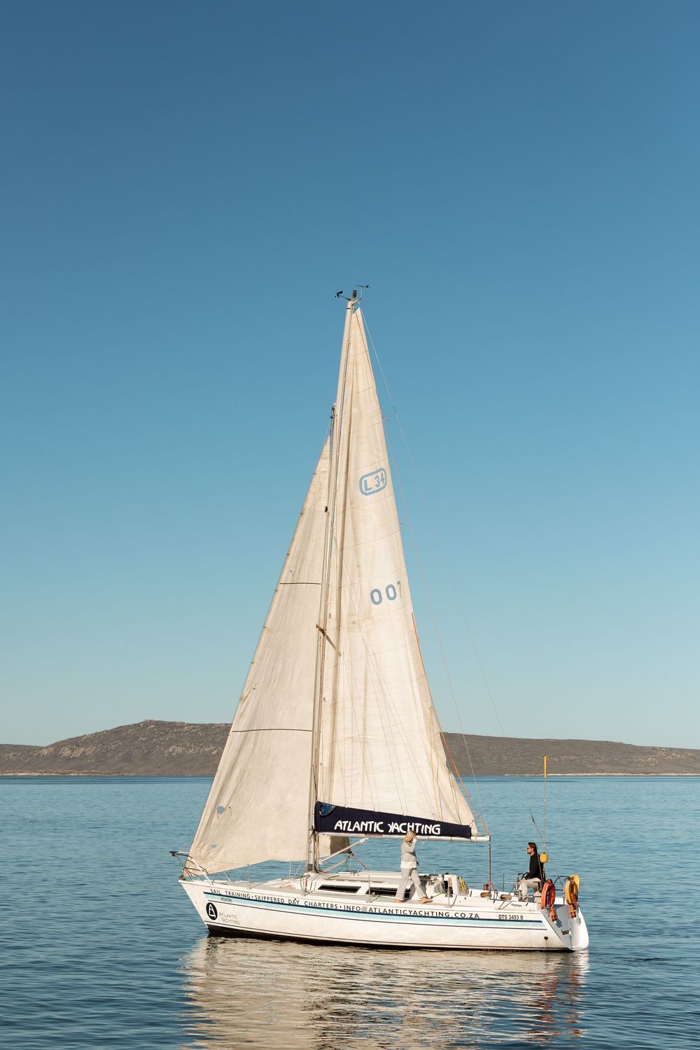 White sailboat with raised sail gliding across calm blue waters near a rocky coastline.