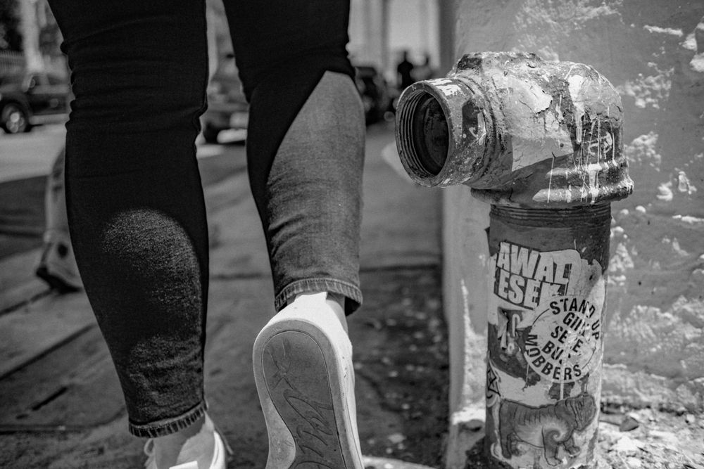 Black and white close-up of legs walking past a graffitied fire hydrant.